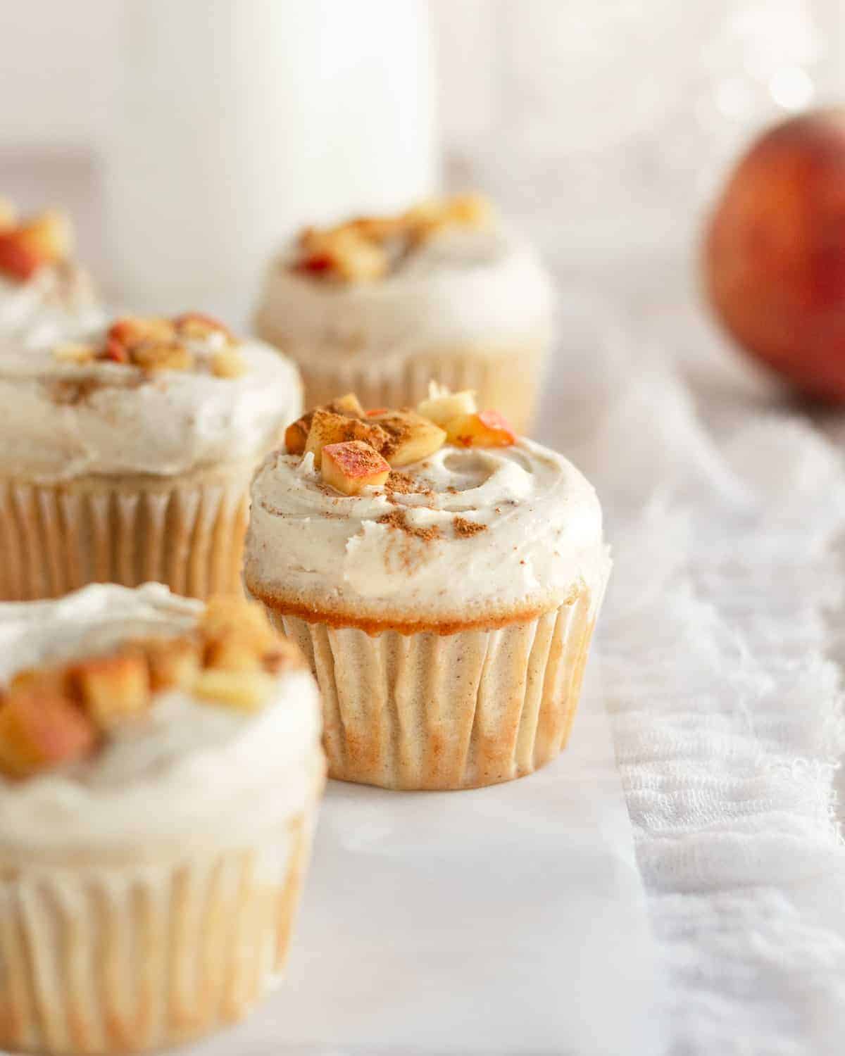 line of frosted peach cupcakes with milk jug and peach behind them.