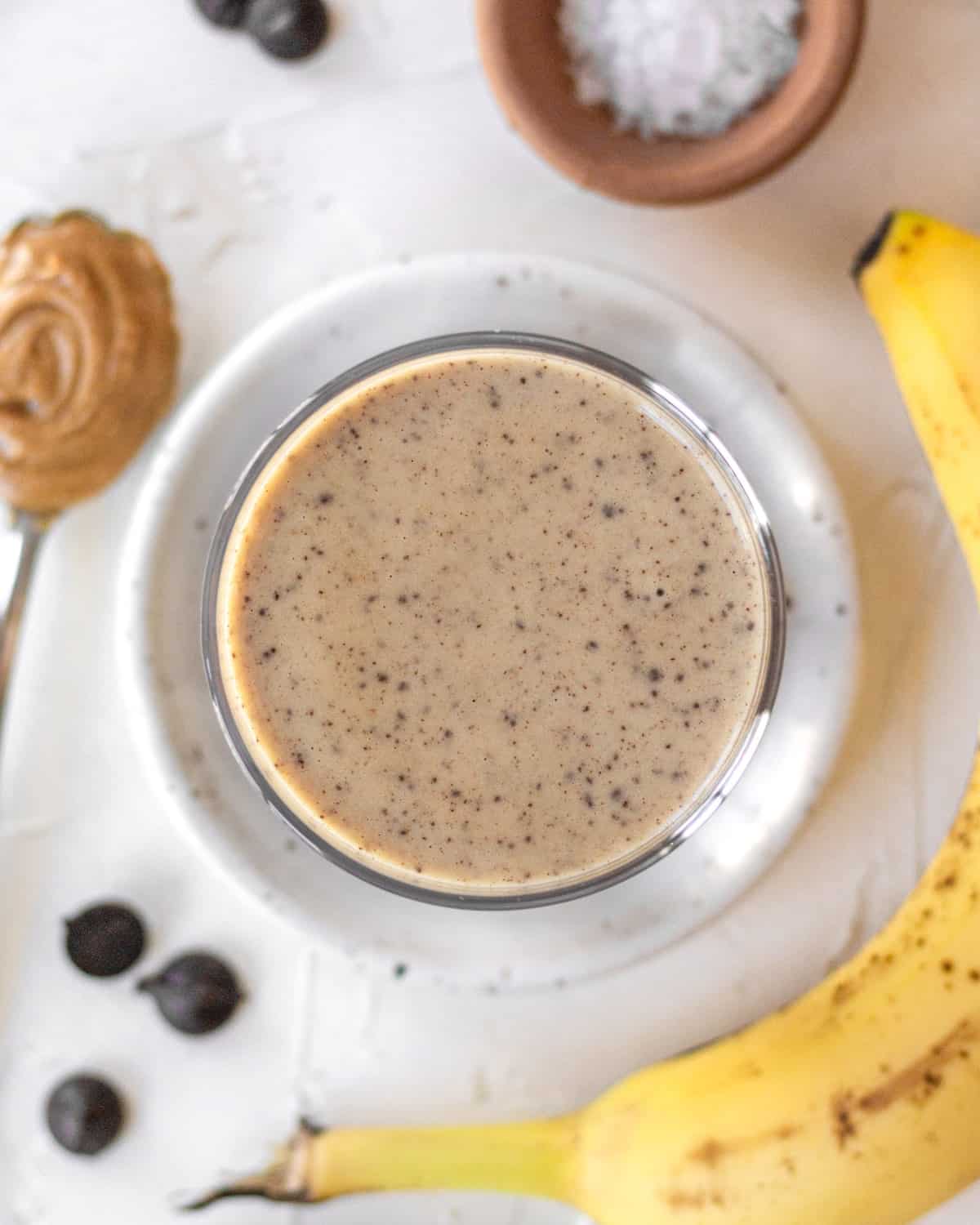 overhead shot of finished smoothie in glass with banana, chocolate chips, salt, and a spoon of peanut butter surrounding it. 
