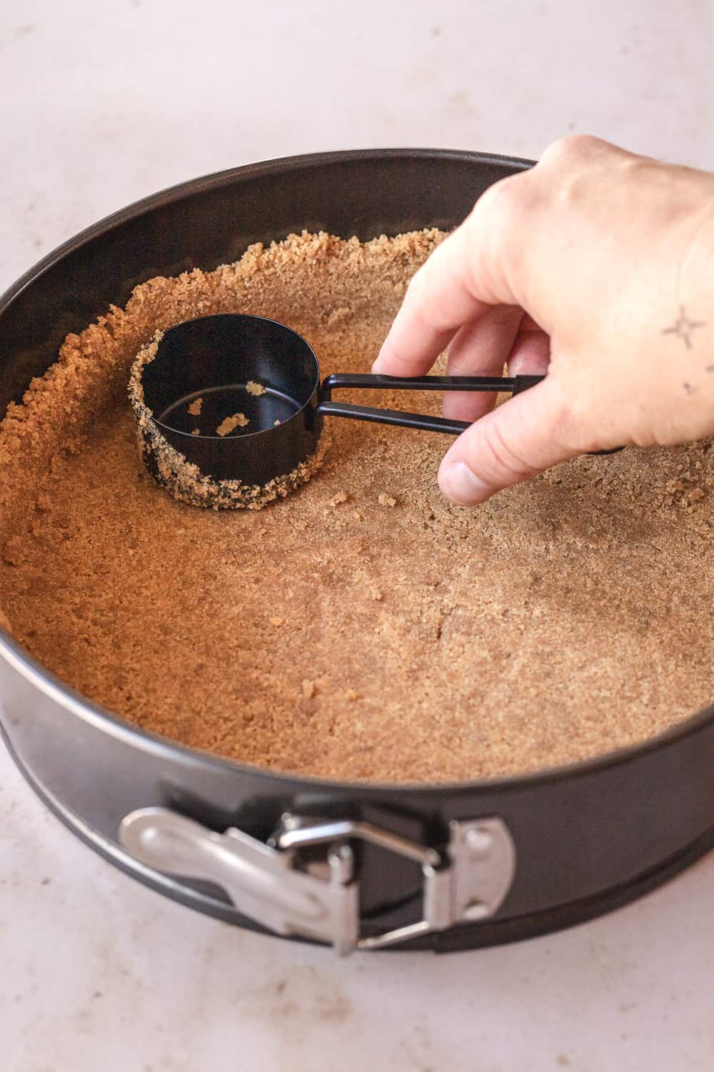graham cracker crust in the springform pan with a measuring cup pushing the crust up the sides of the pan