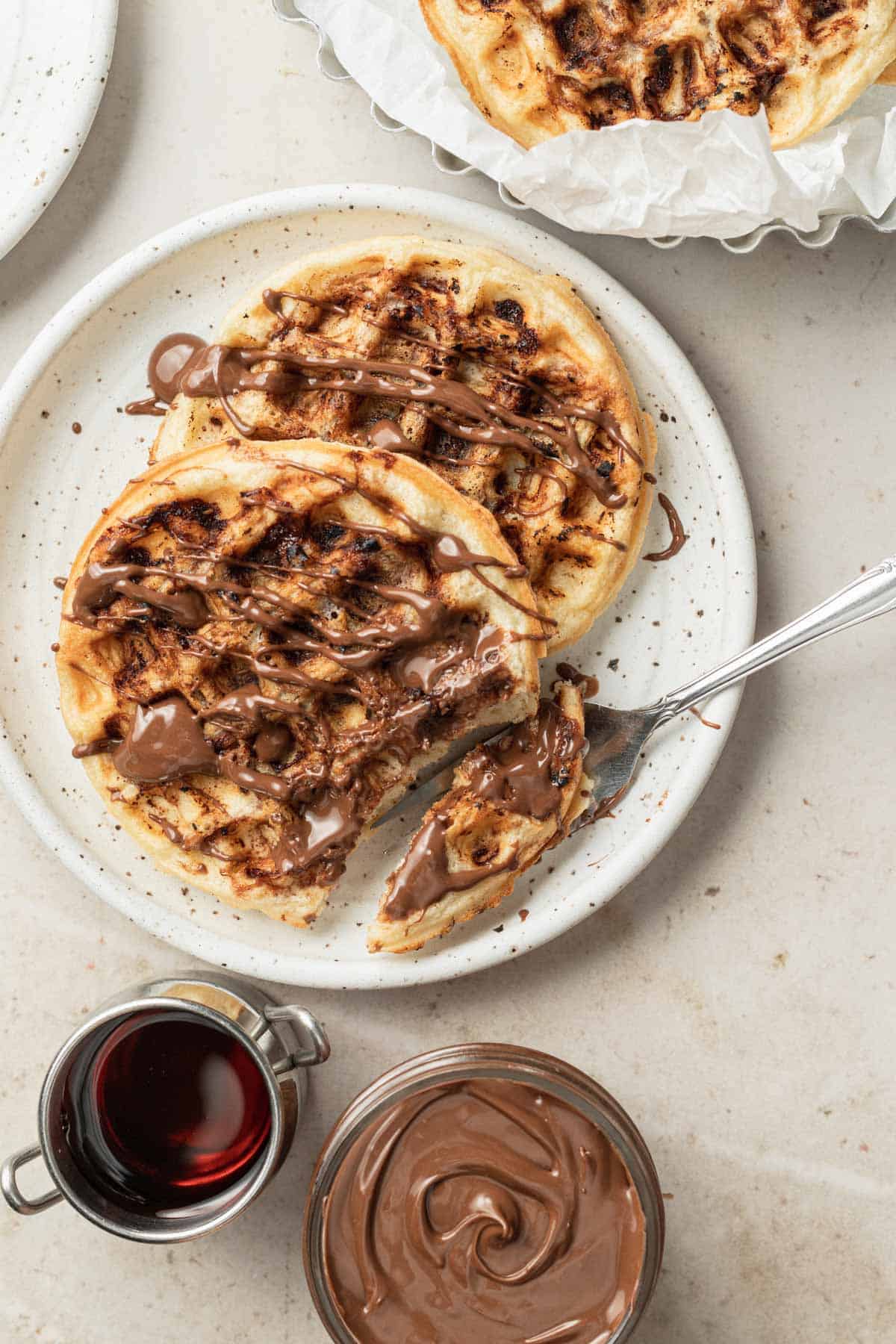 fork taking a bite of waffle from a white plate next to syrup and nutella