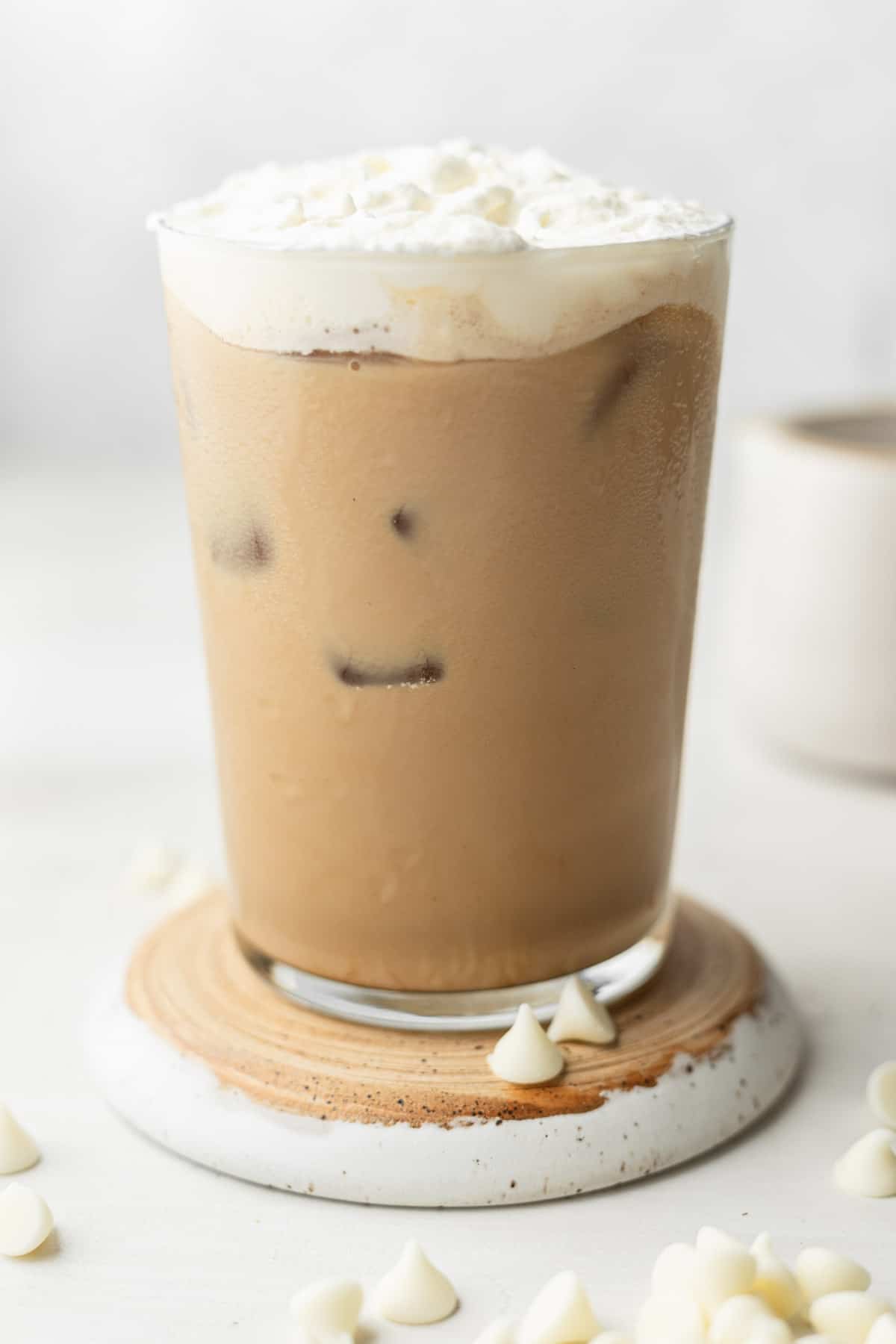 ice white chocolate mocha in a clear glass on a white disc surrounded by white chocolate chips