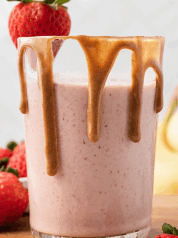 A strawberry peanut butter smoothie in a glass with nut butter dripping down the rim, strawberries and banana in the background.