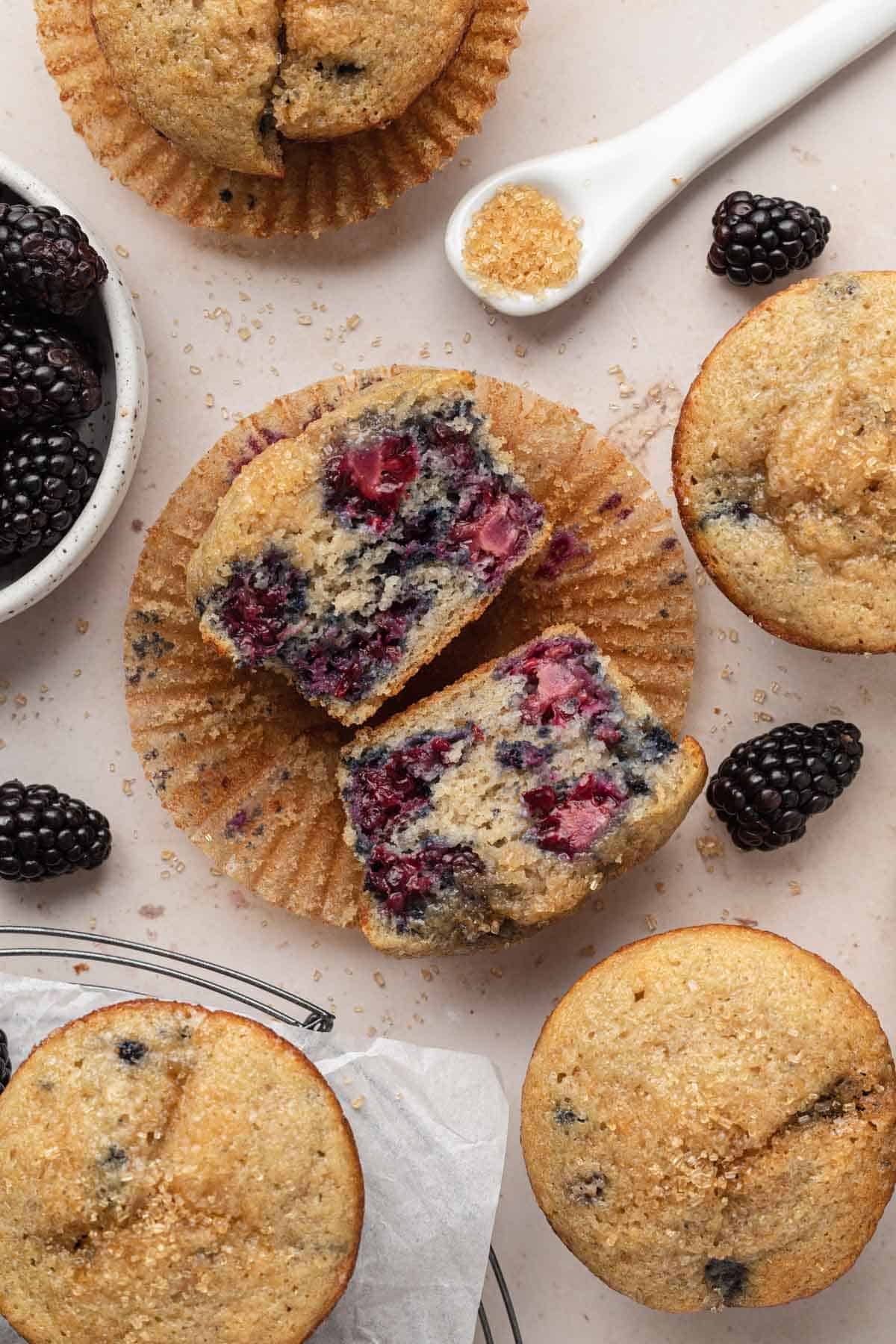 overhead shot of muffin cut in half on a paper liner surround by muffins and blackberries