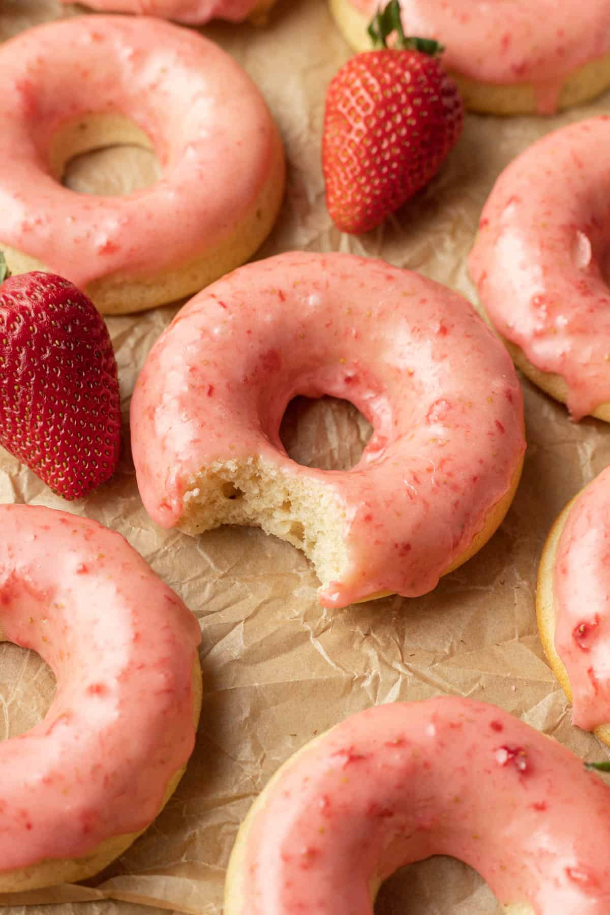 Freshly baked strawberry glazed donuts spread out on parchment paper with one donut in the middle having a bite taken out of it.