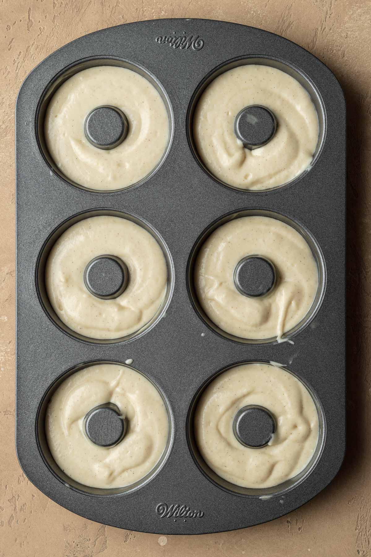 Six donut-shaped wells in a baking pan filled with raw donut batter.