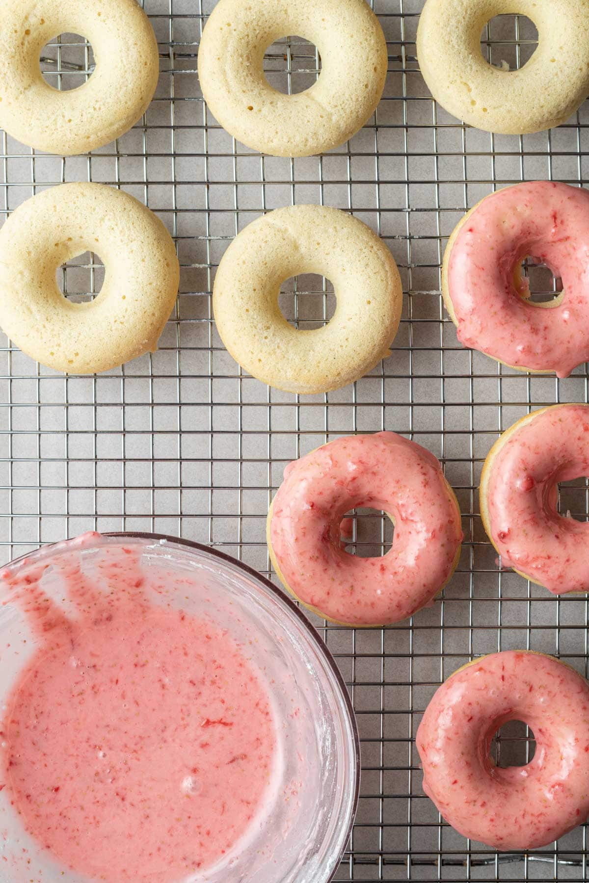 Plain and strawberry glazed donuts on a wire cooling rack next to a bowl of pink glaze.