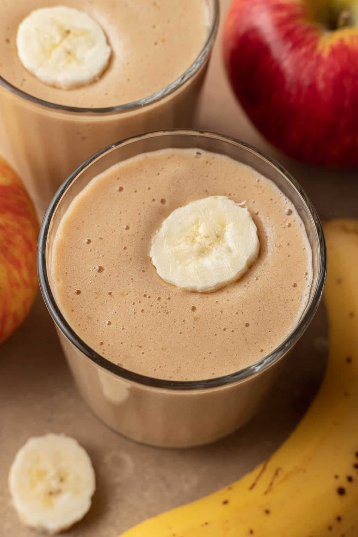 close up view of apple banana smoothie in a clear glass with a slice of banana on top. an apple and a banana are sitting next to the smoothie on a tan backdrop.