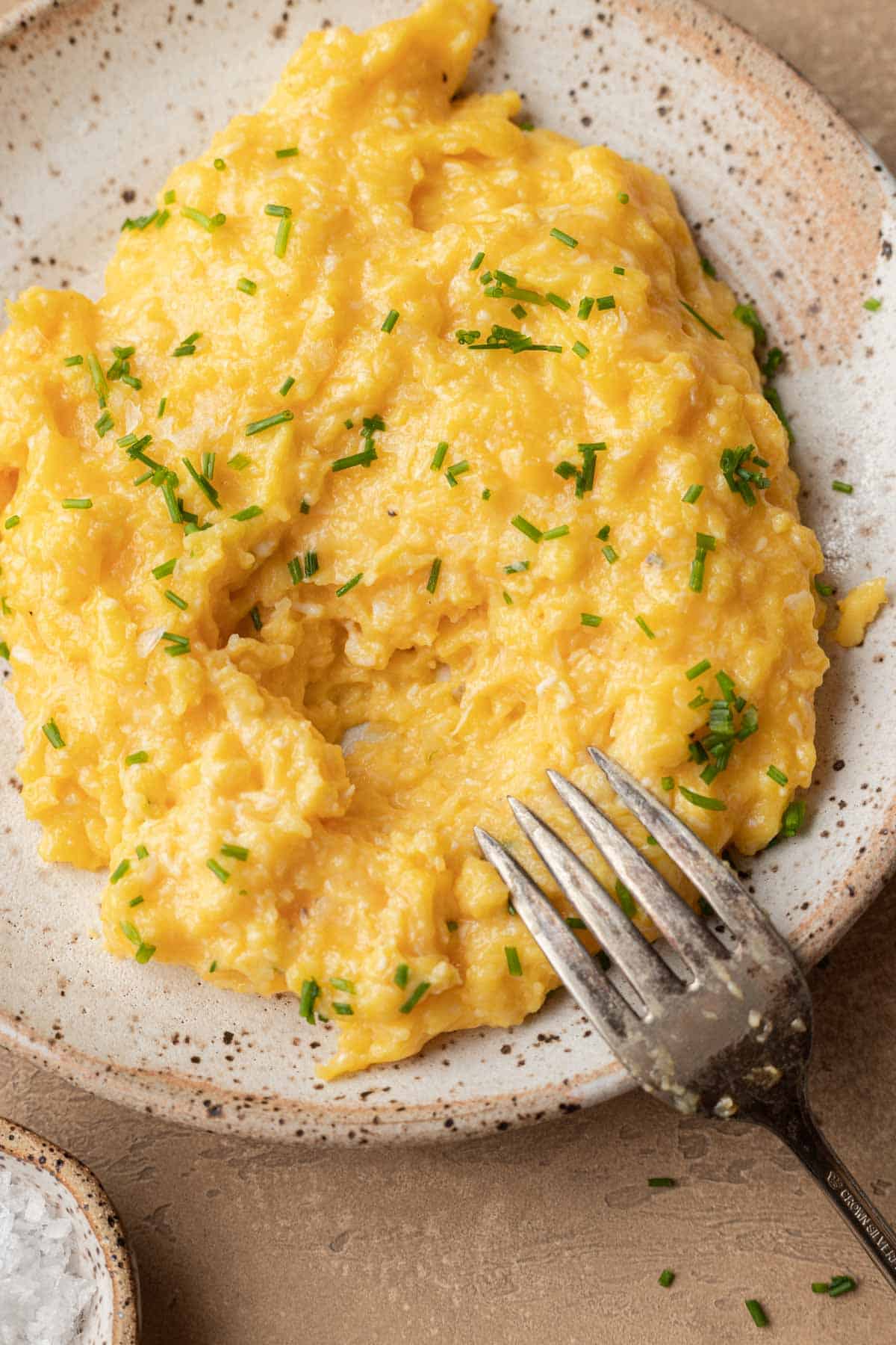 Plate of freshly made cheesy scrambled eggs with chives sprinkled on top and a fork prepped for your first bite.
