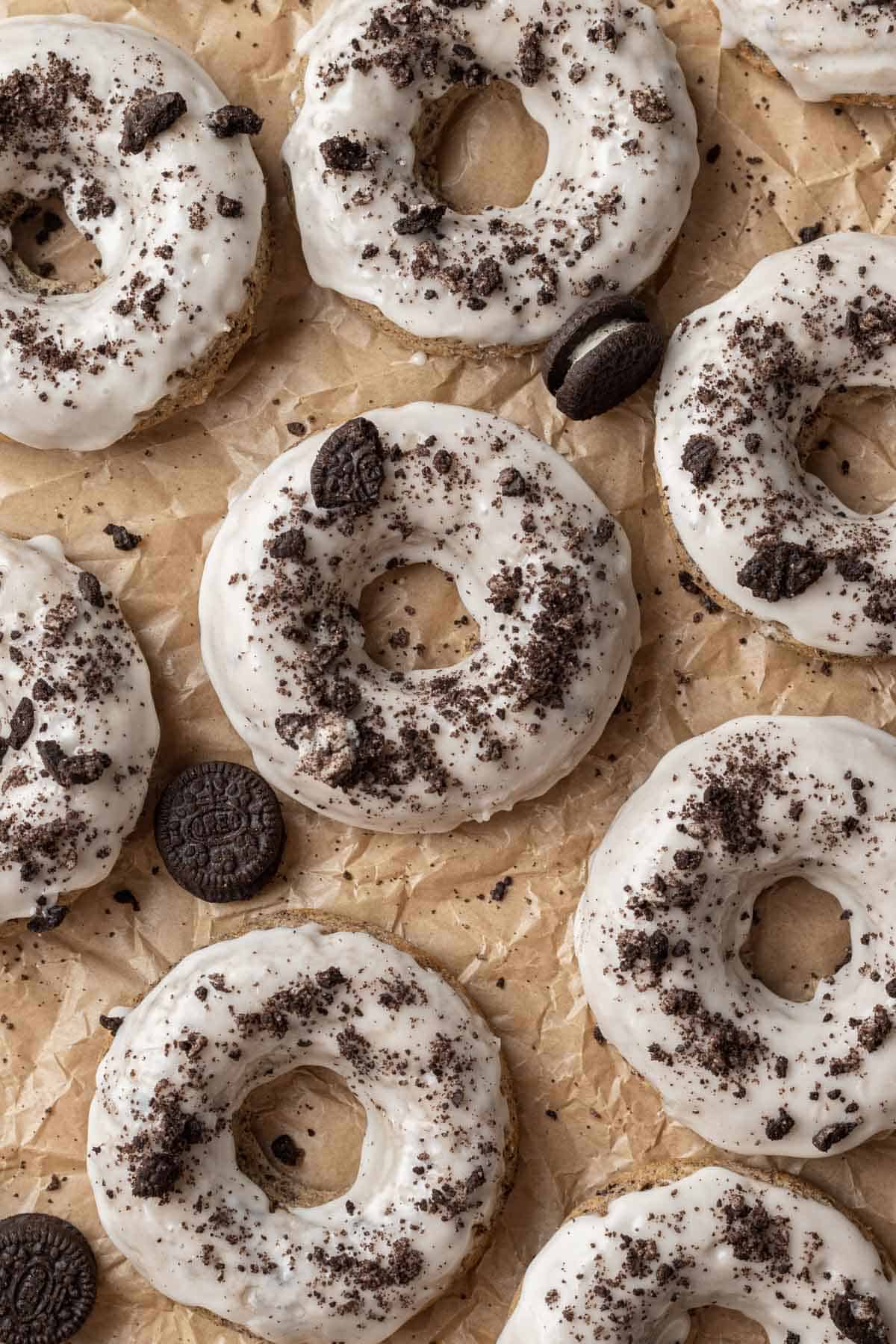 Overhead shot of sheet of freshly baked oreo donuts laying on parchment paper that were just glazed with vanilla icing and topped with cookie crumble.