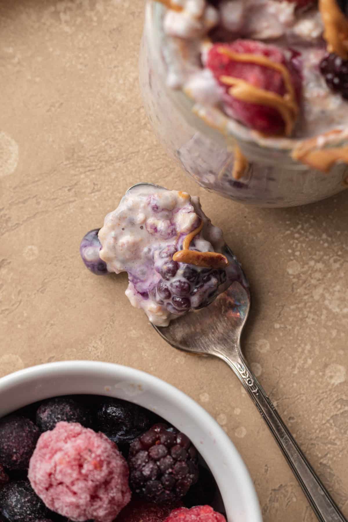 Spoonful of overnight oats with frozen fruit laying on kitchen counter top with glass jar of overnight oats sitting next to it.