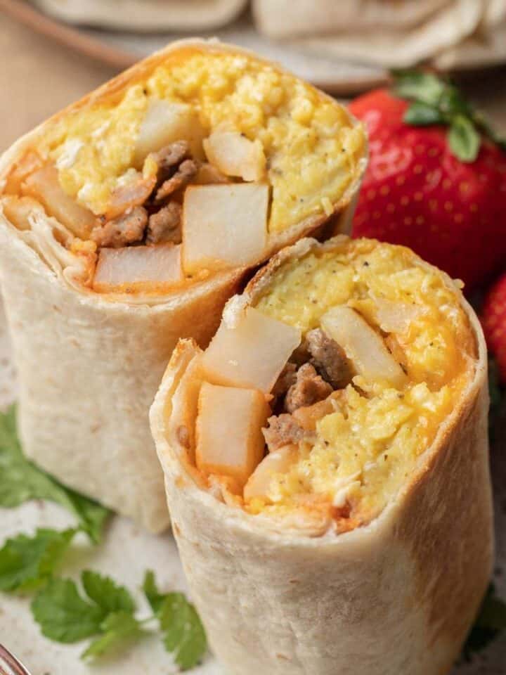 a potato breakfast burrito cut in half on a white plate with strawberries next to it.