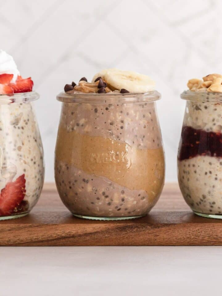 3 jars of different flavored overnight oats on a wooden board with toppings on each.