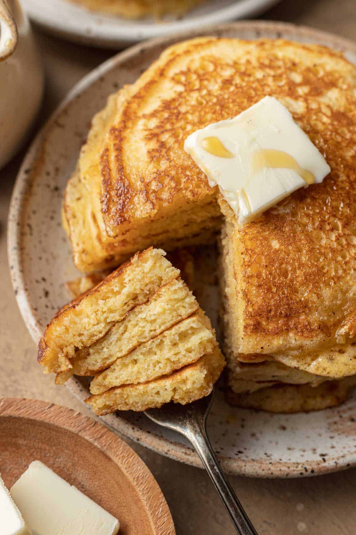 Stack of cornbread pancakes with a fork cutting into the stack. Topped with butter and syrup.