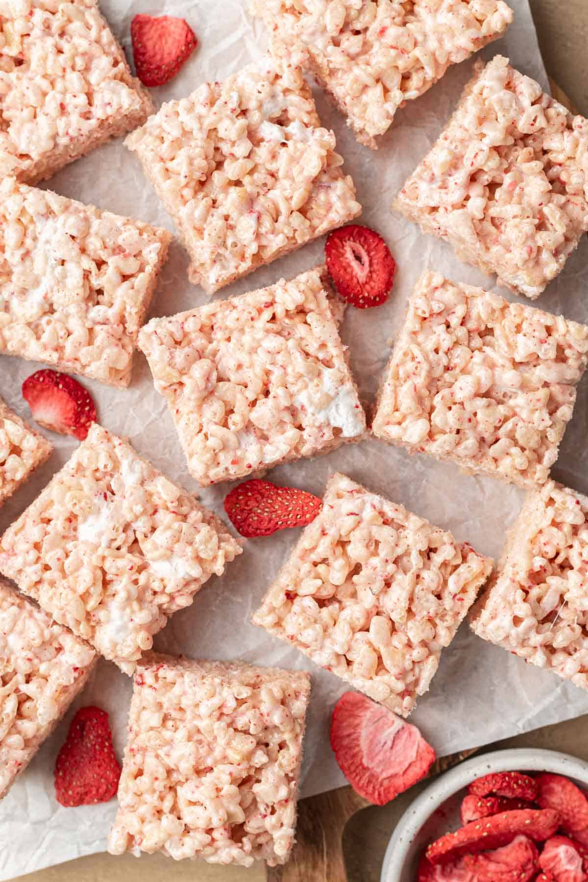 freshly made strawberry rice krispie treats sitting on parchment paper with freezer dried strawberries surrounding them.