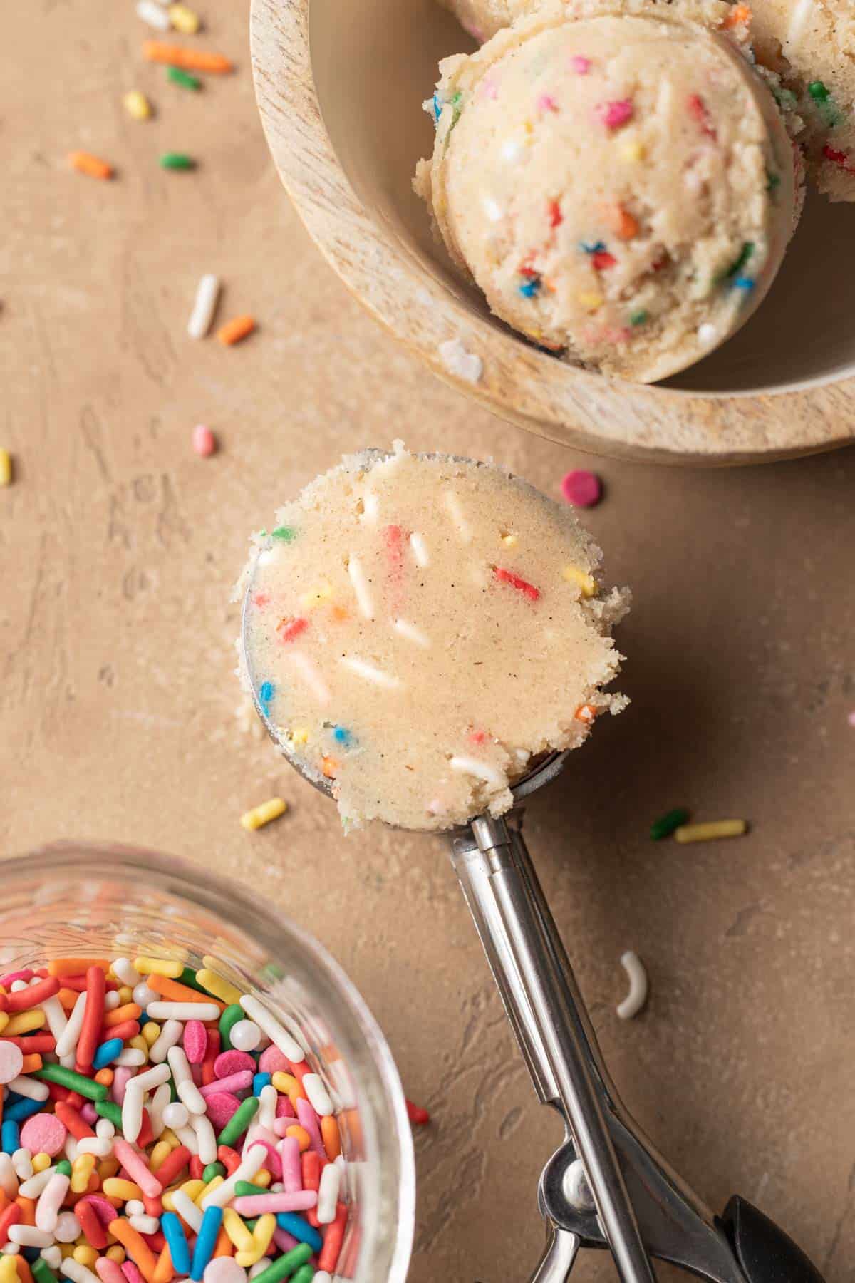 Scoop of edible sugar cookie dough that is still in the cookie scooper next to a bowl of sprinkles.