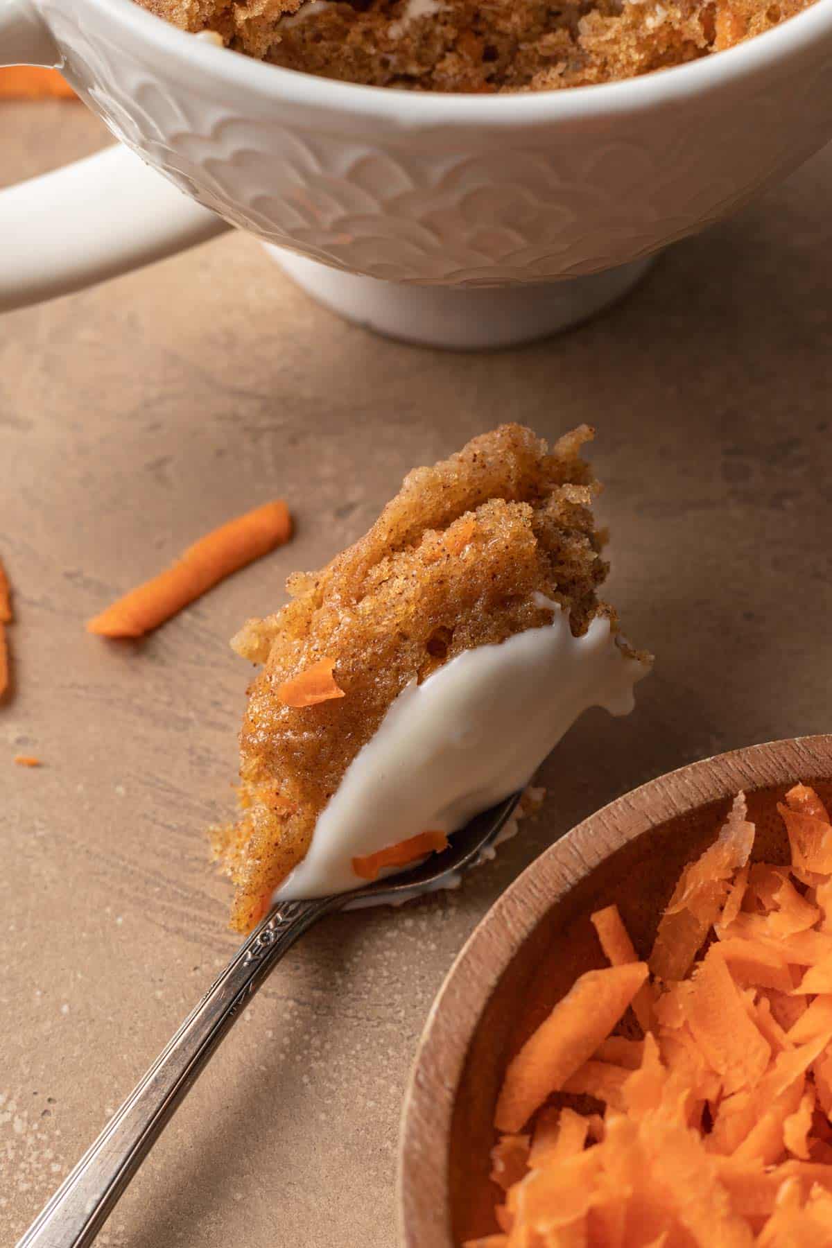 Big spoonful of carrot cake mug cake with frosting on top sitting next to a bowl of shredded carrots.