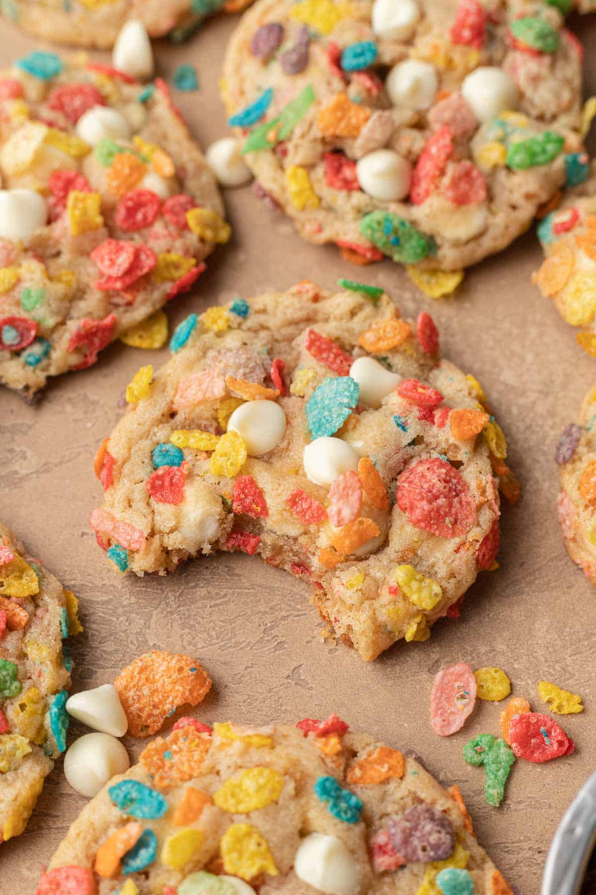 fruity pebble cookie on a tan backdrop with a bite taken out of it, surrounded by other cookies