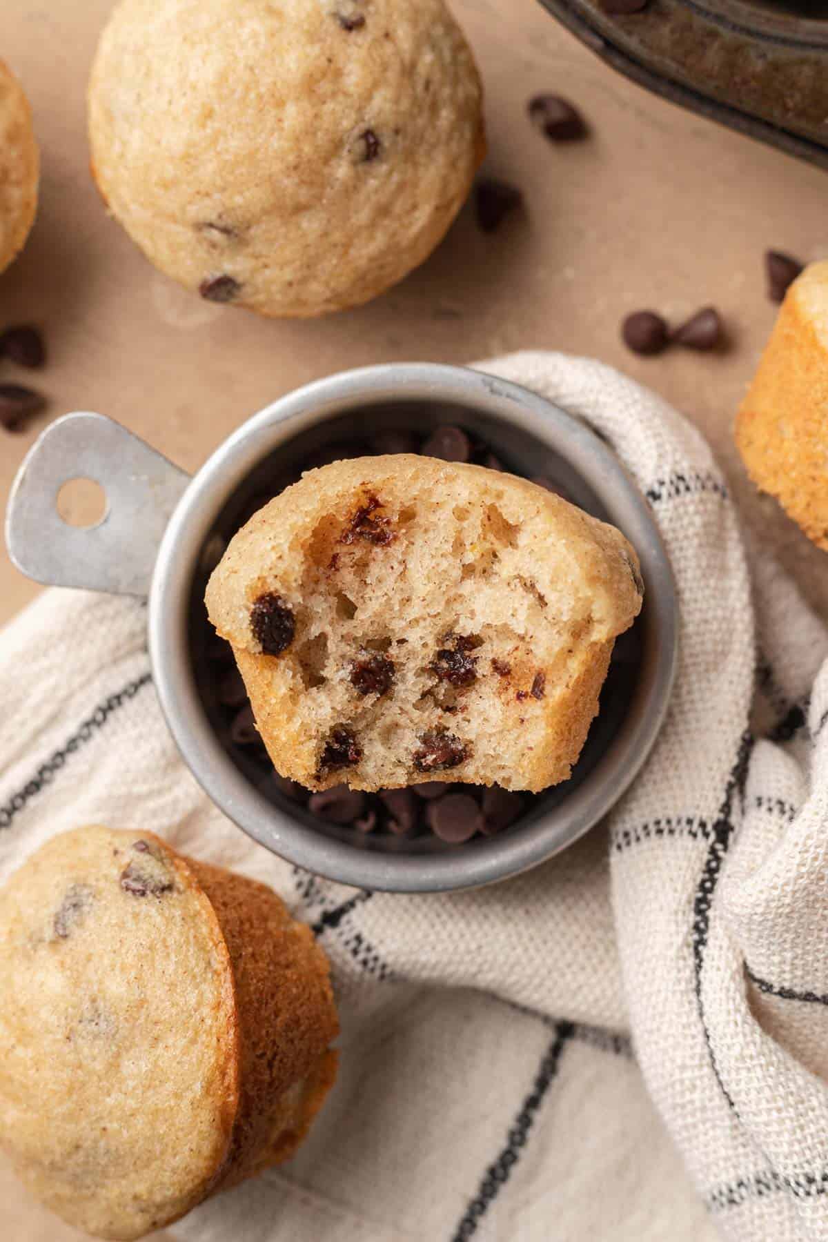 mini chocolate chip muffin with a bite taken out of it laying in a bowl of mini chocolate chips with muffins around it.