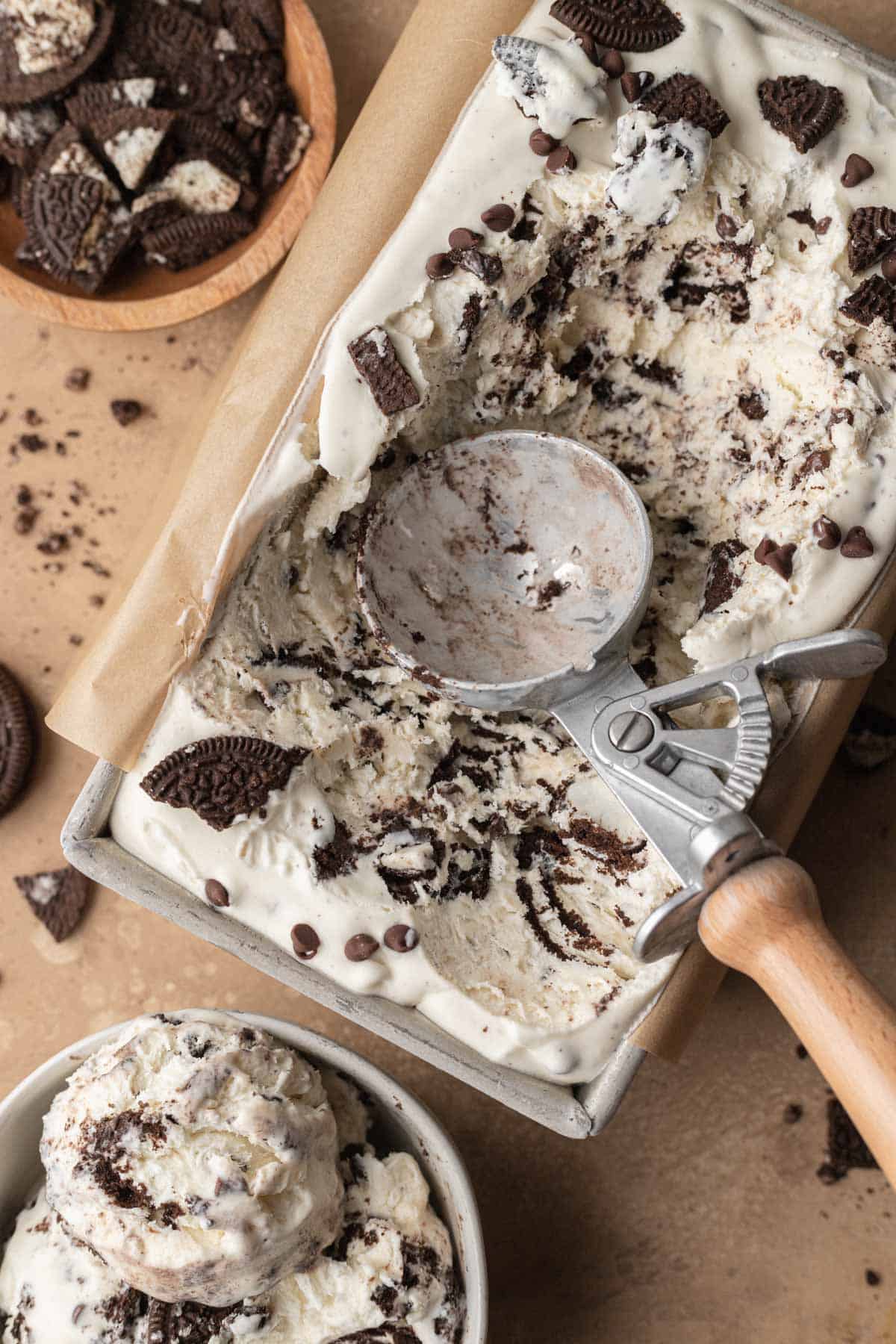 Oreo ice cream in a rectangle dish with a metal ice cream scoop on top, with a bowl of scooped ice cream next to it.