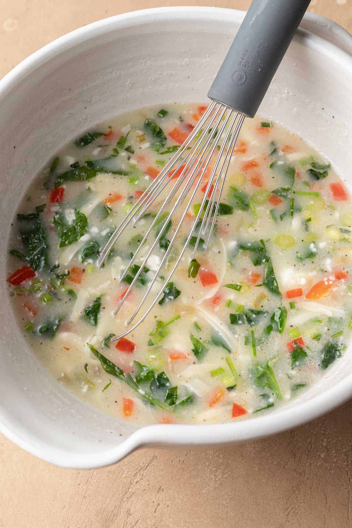 Egg whites with vegetable mix ins in a white glass mixing bowl with a whisk.