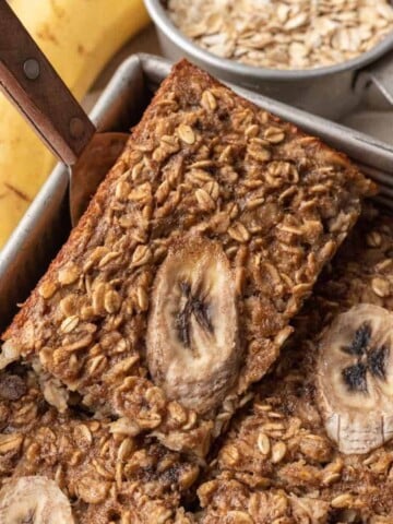 A piece of banana bread baked oats being scooped out of a tin baking pan by a spatula.