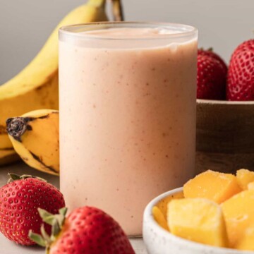Mango strawberry banana smoothie in glass cup with ingredients sitting beside it.