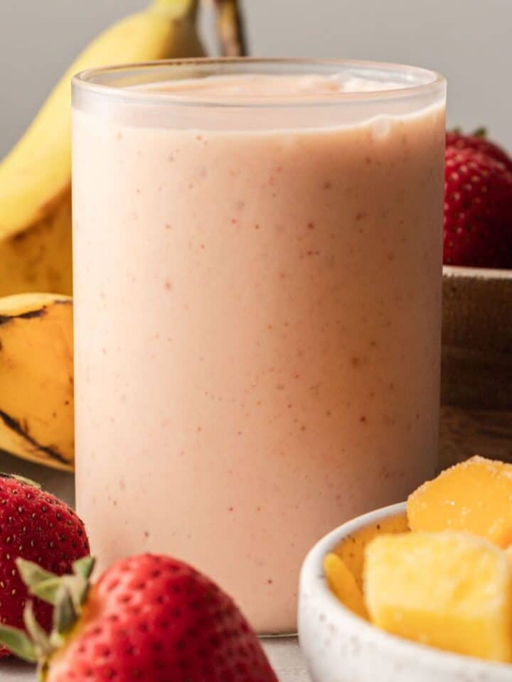 Mango strawberry banana smoothie in glass cup with ingredients sitting beside it.