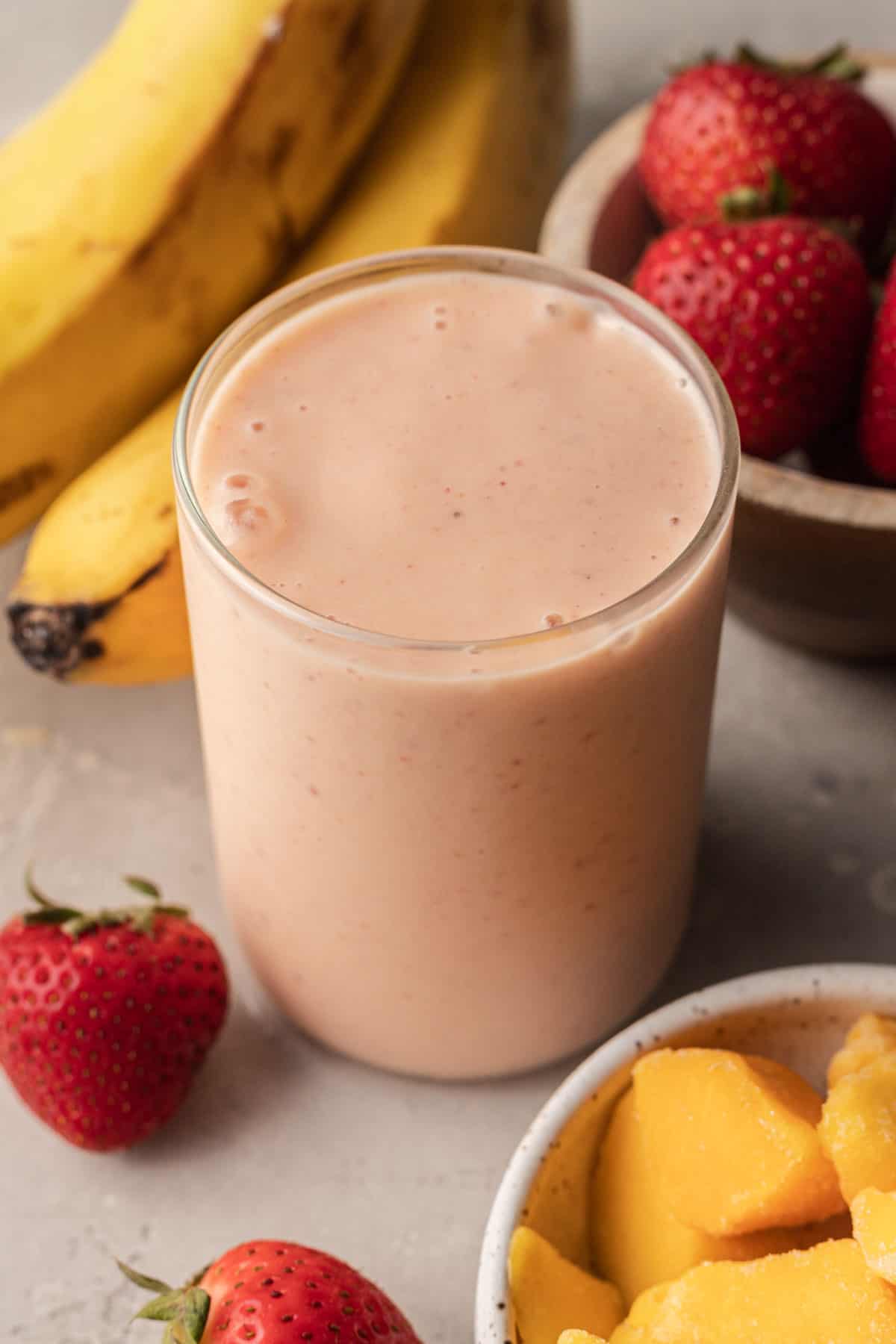Mango strawberry banana smoothie in clear glass with fruit ingredients placed around it.