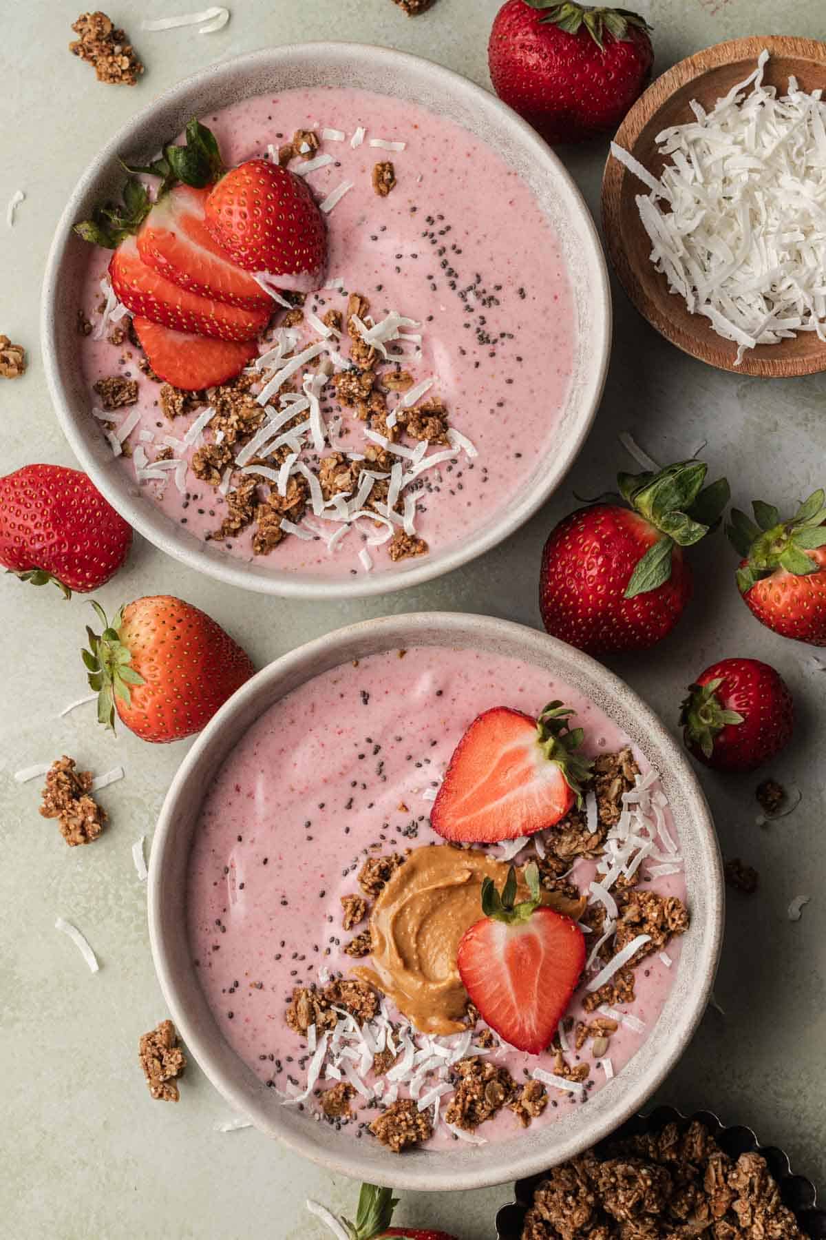 Two strawberry smoothie bowls in ceramic bowls topped with fresh strawberries, coconut flakes and granola.