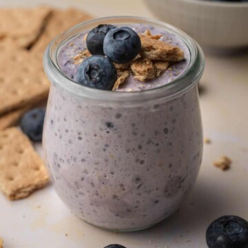 blueberry cheesecake overnight oats in a glass jar topped with blueberries and graham crackers