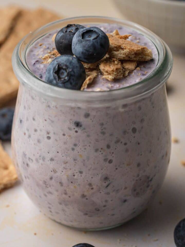 blueberry cheesecake overnight oats in a glass jar topped with blueberries and graham crackers