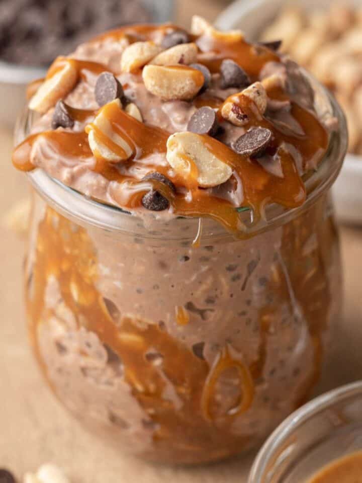 snickers overnight oats in a class jar topped with peanuts, chocolate chips, and a drizzle of caramel.