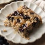 a slice of blueberry baked oatmeal on a scalloped plate with maple syrup drizzled on top.