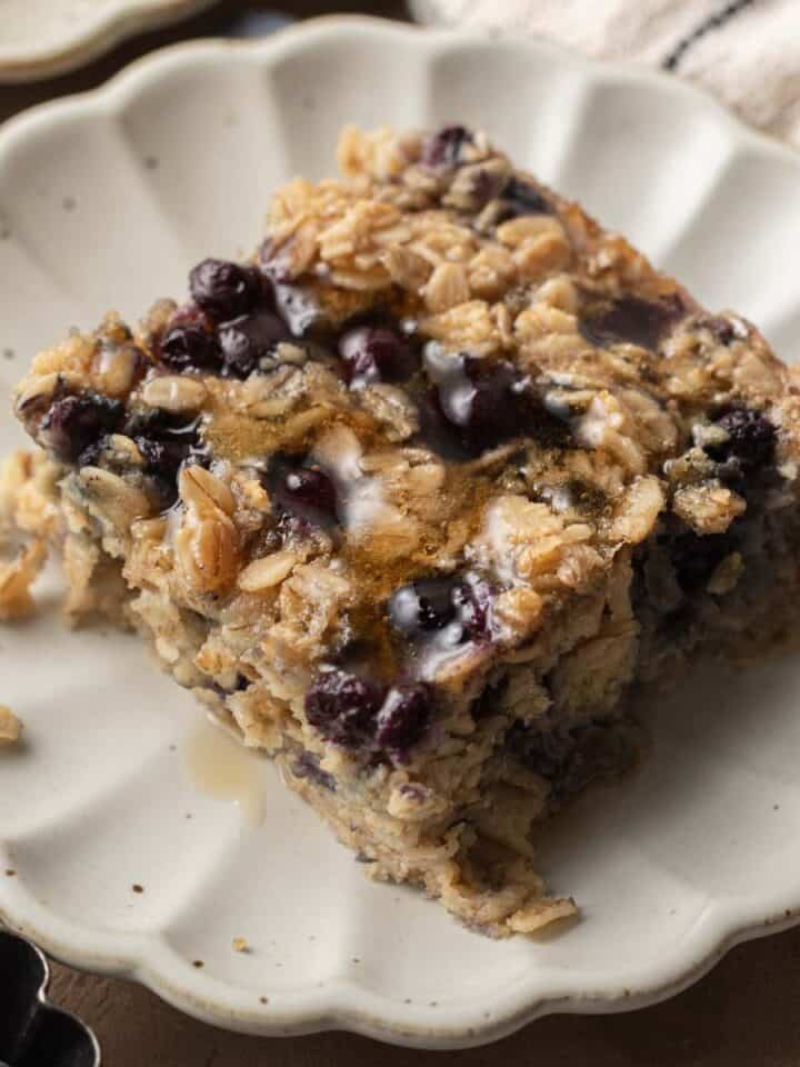 a slice of blueberry baked oatmeal on a scalloped plate with maple syrup drizzled on top.