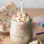 birthday cake overnight oats in a glass jar topped with whipped cream, sprinkles, and a candle, with a slice of birthday cake behind it.