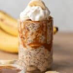 Glass of caramel banana overnight oats topped with whipped cream, evoking the delightful flavors of banoffee pie, with ripe bananas in the background.