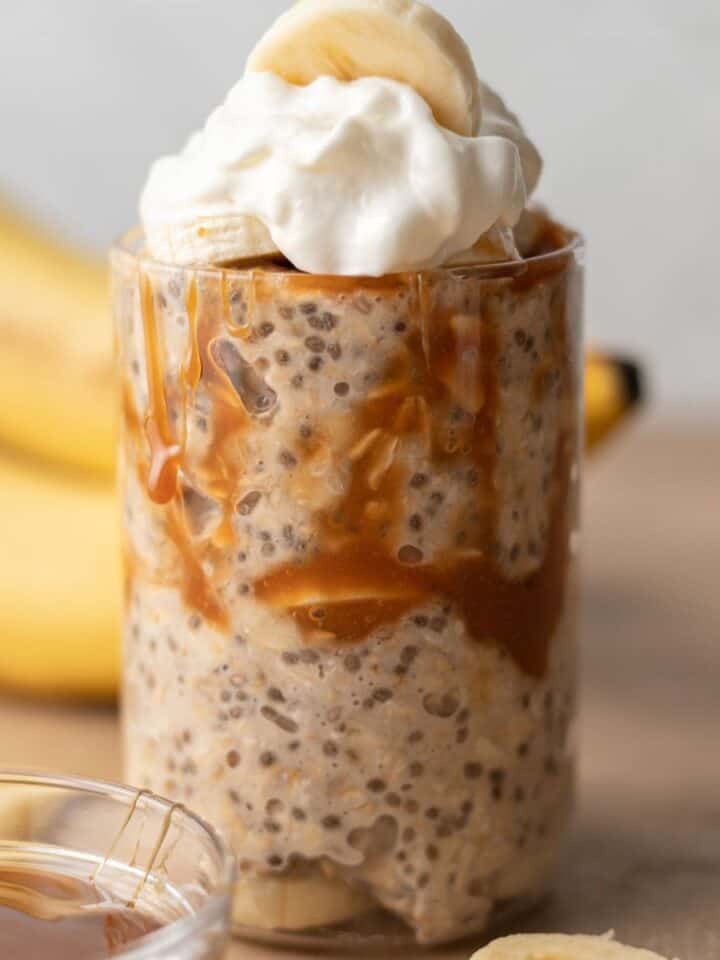 Glass of caramel banana overnight oats topped with whipped cream, evoking the delightful flavors of banoffee pie, with ripe bananas in the background.