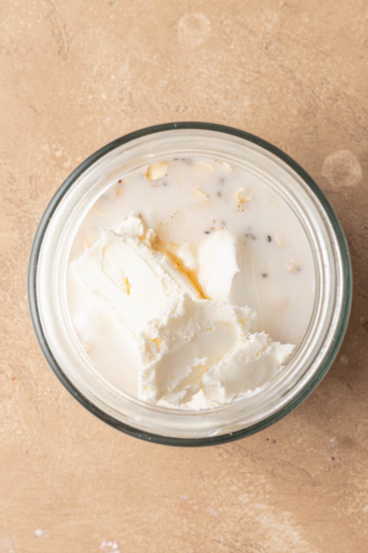 A glass jar filled with oat mixture and liquid ingredients, including cream cheese, on a beige surface.