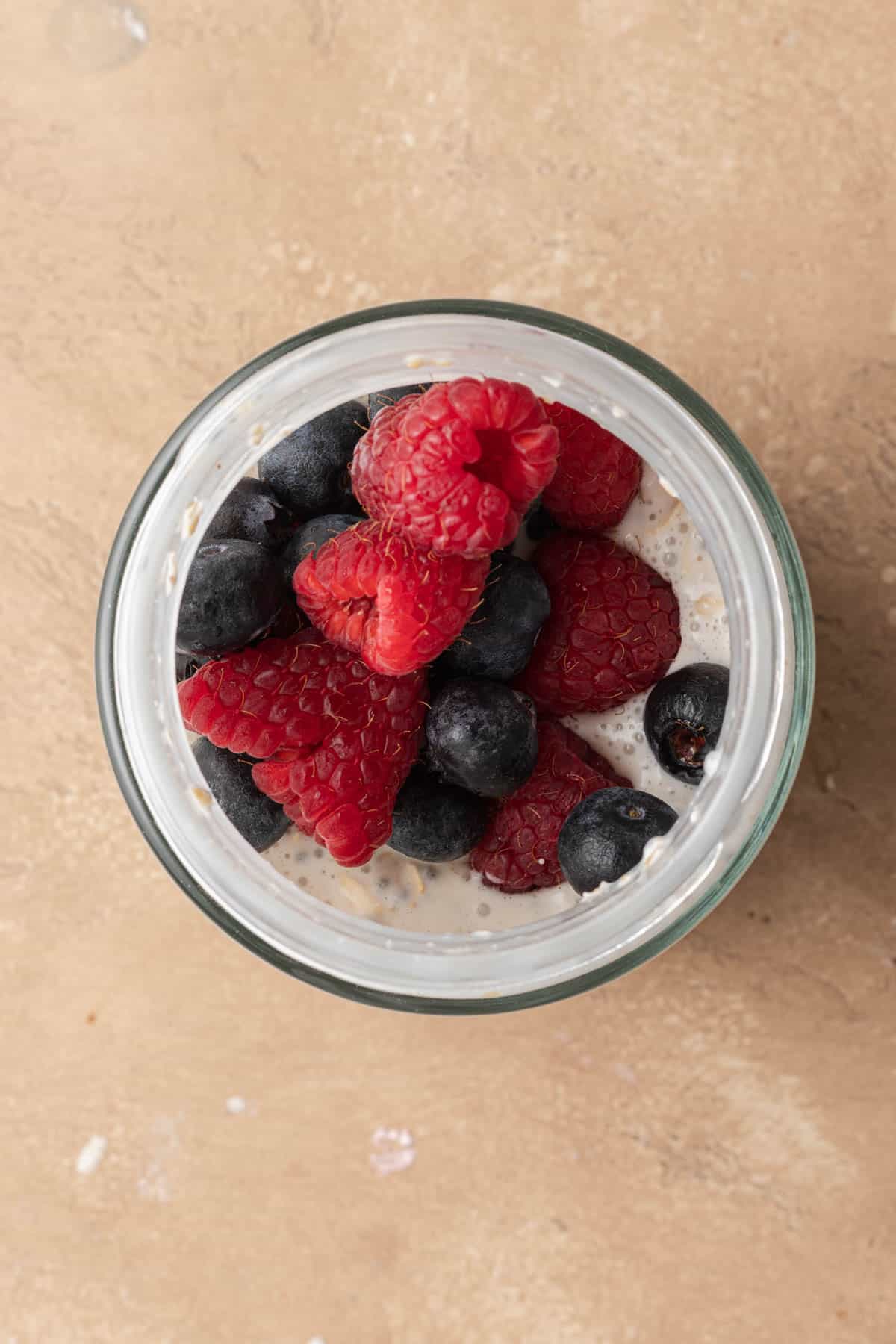 A glass jar filled with mixed oatmeal, topped with fresh raspberries and blueberries, ready to sit in the fridge overnight.