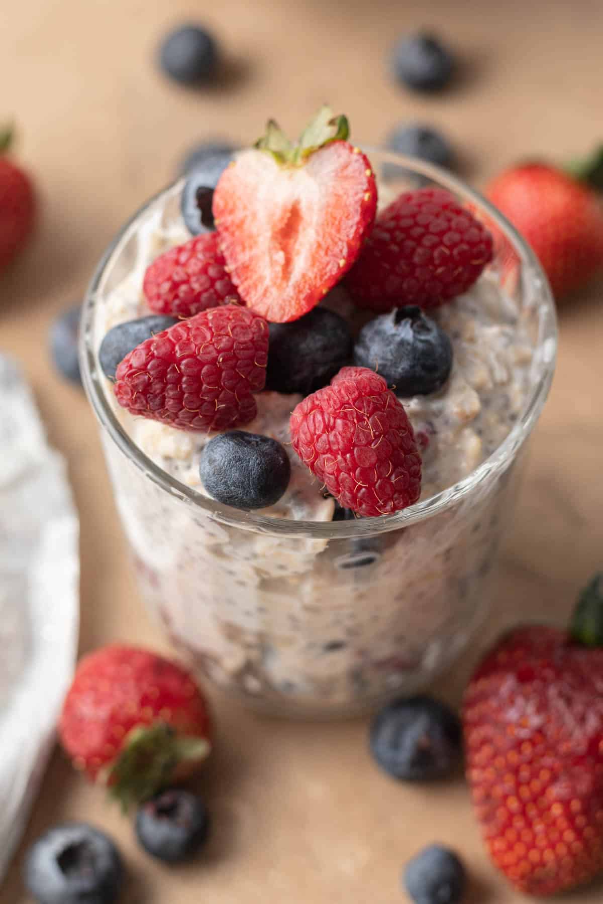 Glass of berry cheesecake overnight oatmeal topped with sliced strawberries, raspberries, and blueberries, surrounded by more berries.