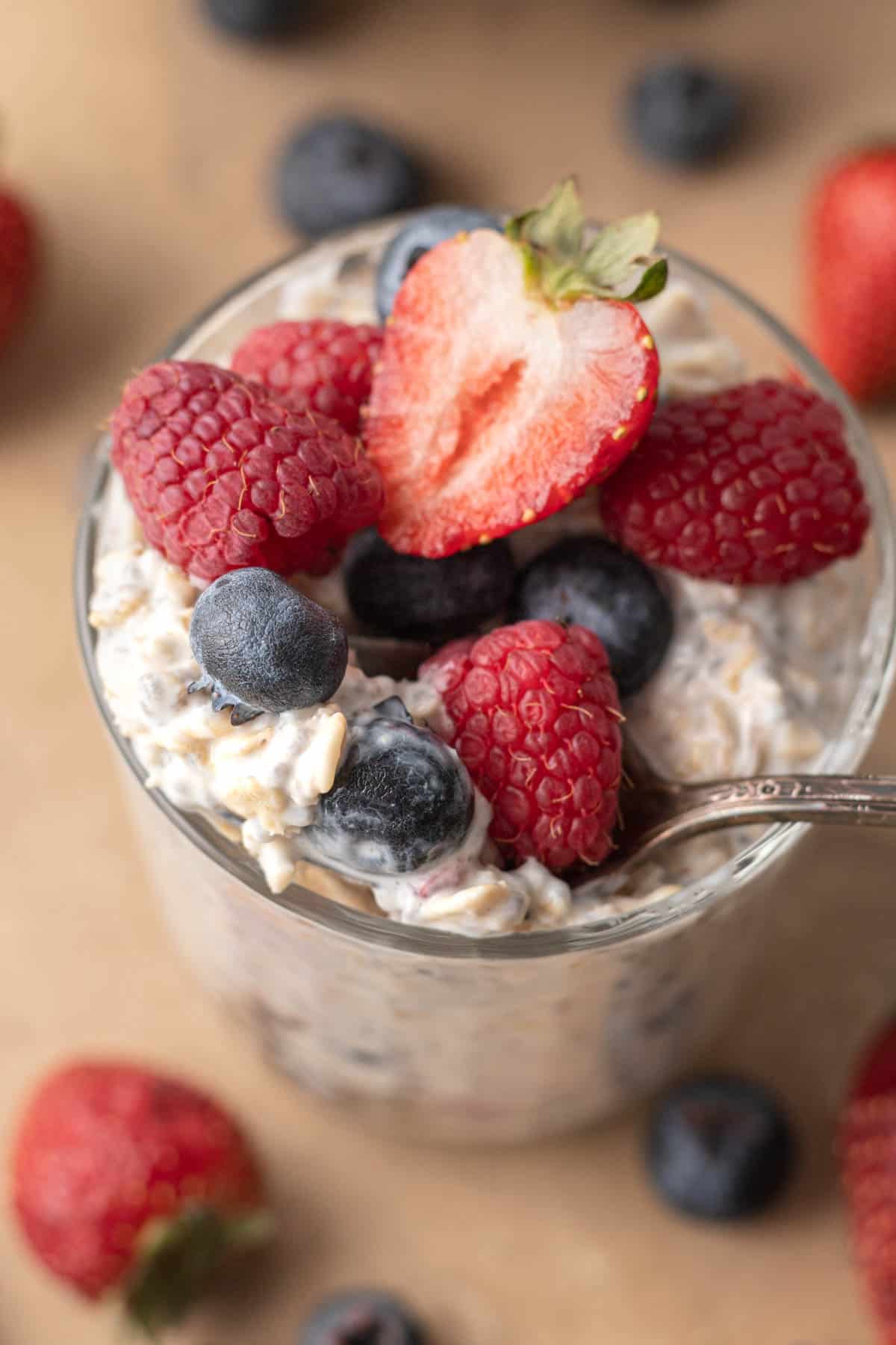 Glass of oatmeal topped with raspberries, blueberries, and a halved strawberry, with more berries scattered around.