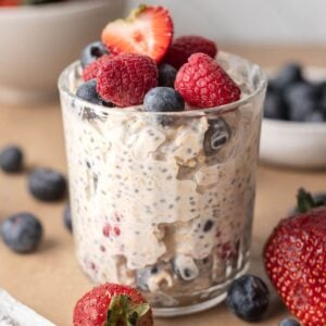 A glass of creamy berry cheesecake overnight oats topped with raspberries, blueberries, and half a strawberry.