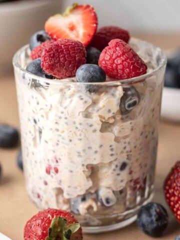A glass of creamy berry cheesecake overnight oats topped with raspberries, blueberries, and half a strawberry.
