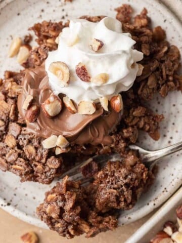 A small plate with nutella baked oatmeal, whipped cream, and chopped nuts, with a fork on the side.