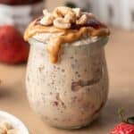 Jar of peanut butter and jelly overnight oats topped with nuts, with strawberries in the background.