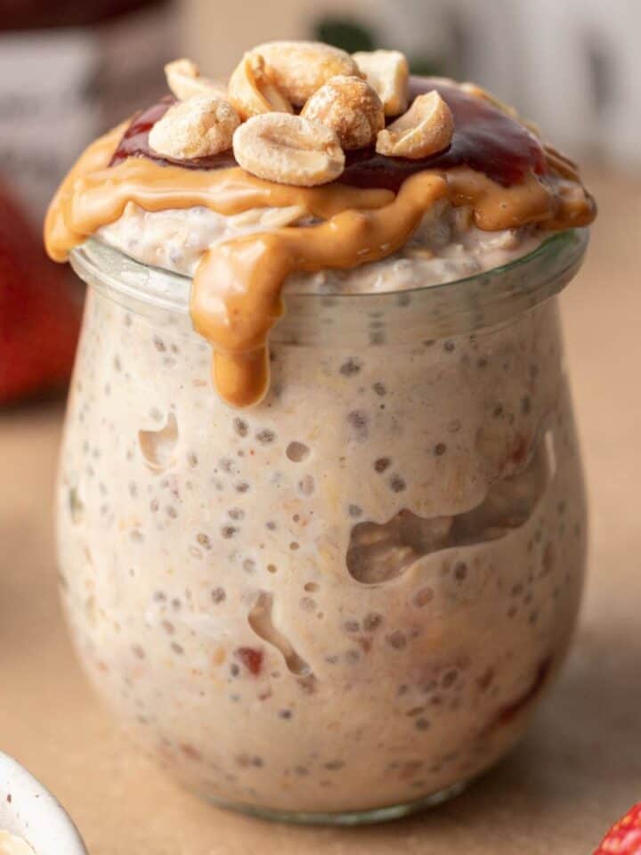 Jar of peanut butter and jelly overnight oats topped with nuts, with strawberries in the background.