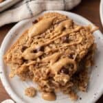 A slice of peanut butter baked oats crowned with chocolate chips rests invitingly on a white plate.