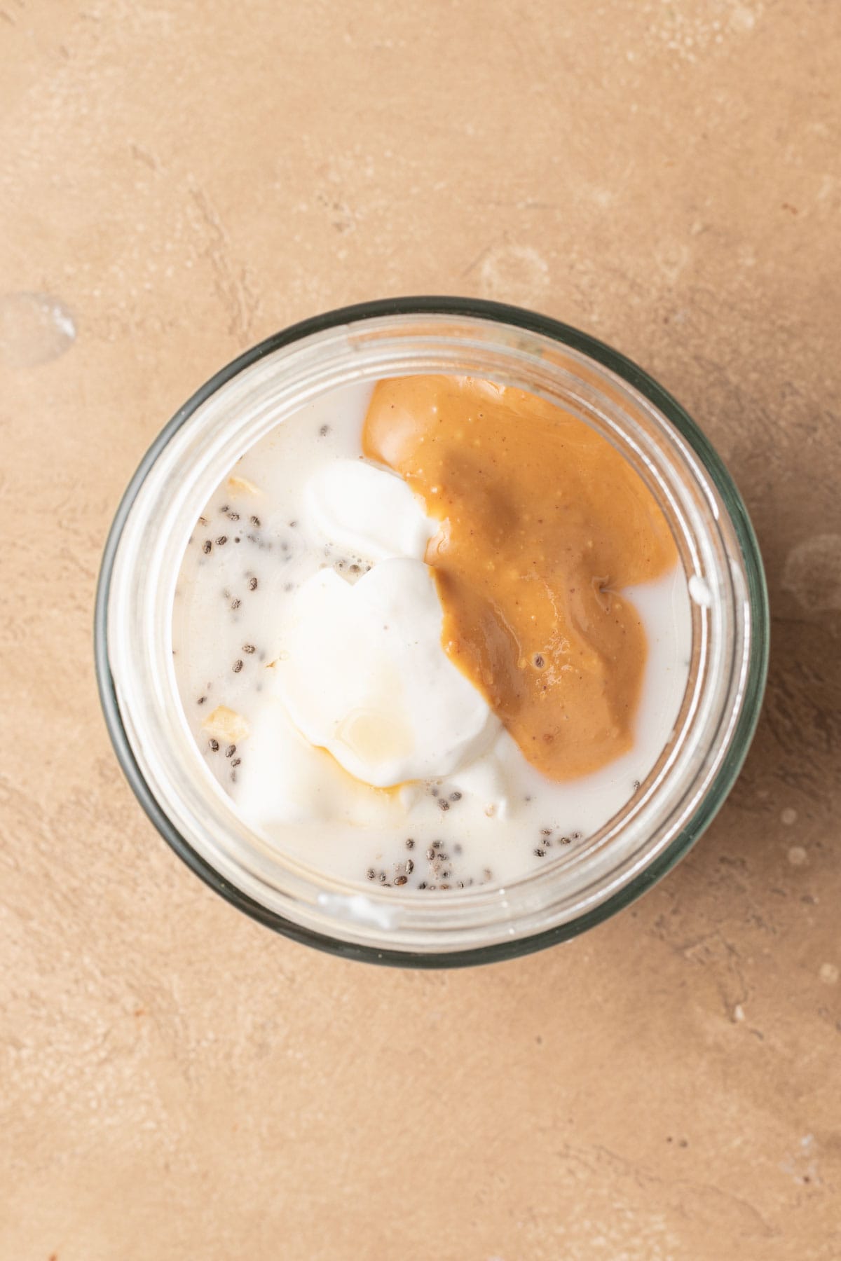 A glass jar with creamy yogurt & milk added, chia seeds and oats, and a swirl of peanut butter sits on a beige surface.