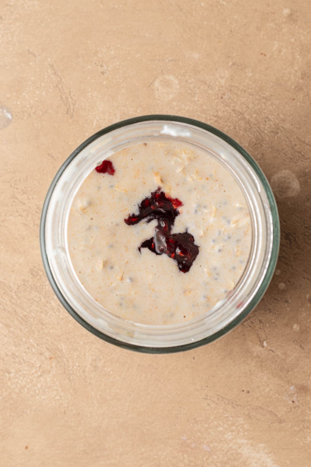 Glass jar of creamy pb&j overnight oats topped with a dollop of dark berry jam on a beige textured surface.