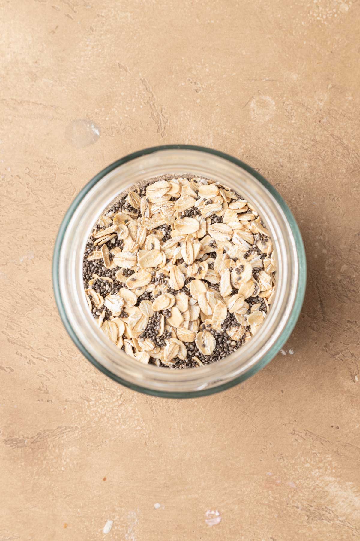 A glass jar filled with pb&j overnight oats, packed with chia seeds, sits on a textured beige surface.